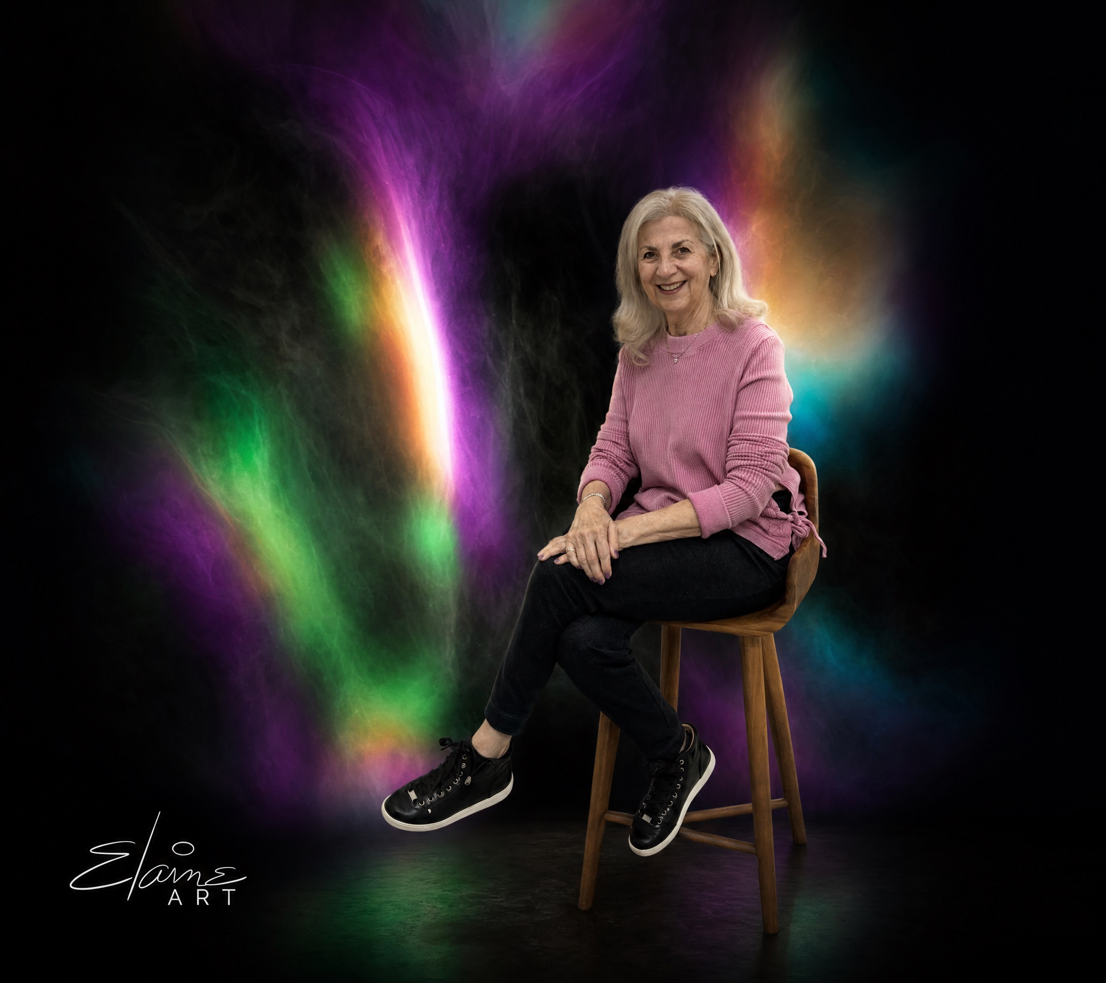 Elaine Clarfield Gitalis seated in her studio before a swirling violet-and-green light painting, Elaine Art signature bottom-left